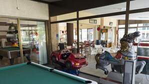 Una mesa de billar, un coche en miniatura y un caballo de carrusel en una habitación con ventanas amplias y una pared de ladrillo.