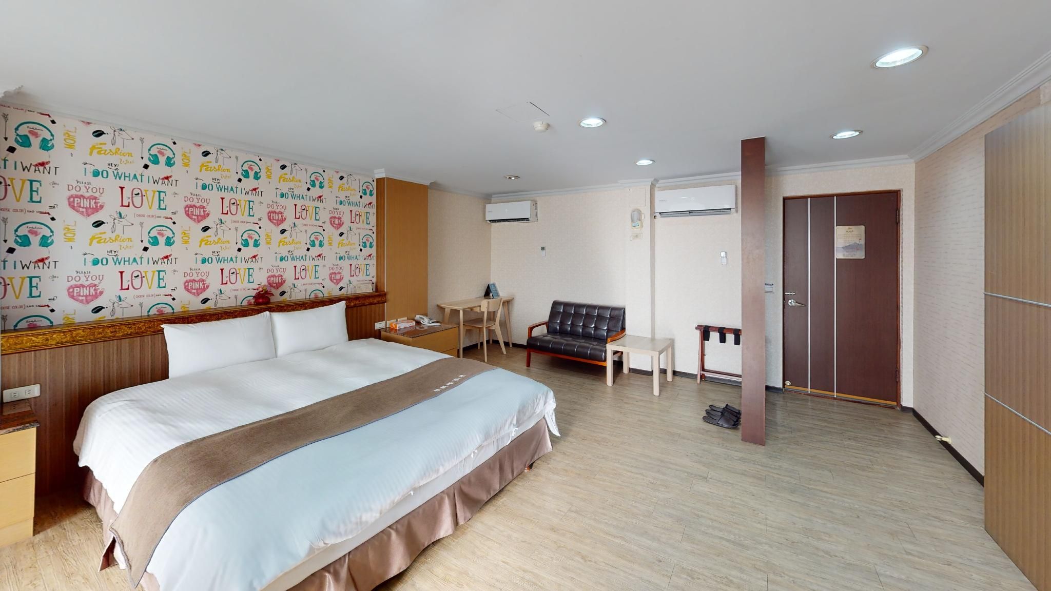 A hotel room with a large bed, a desk, a chair, a sofa, and a wardrobe.
