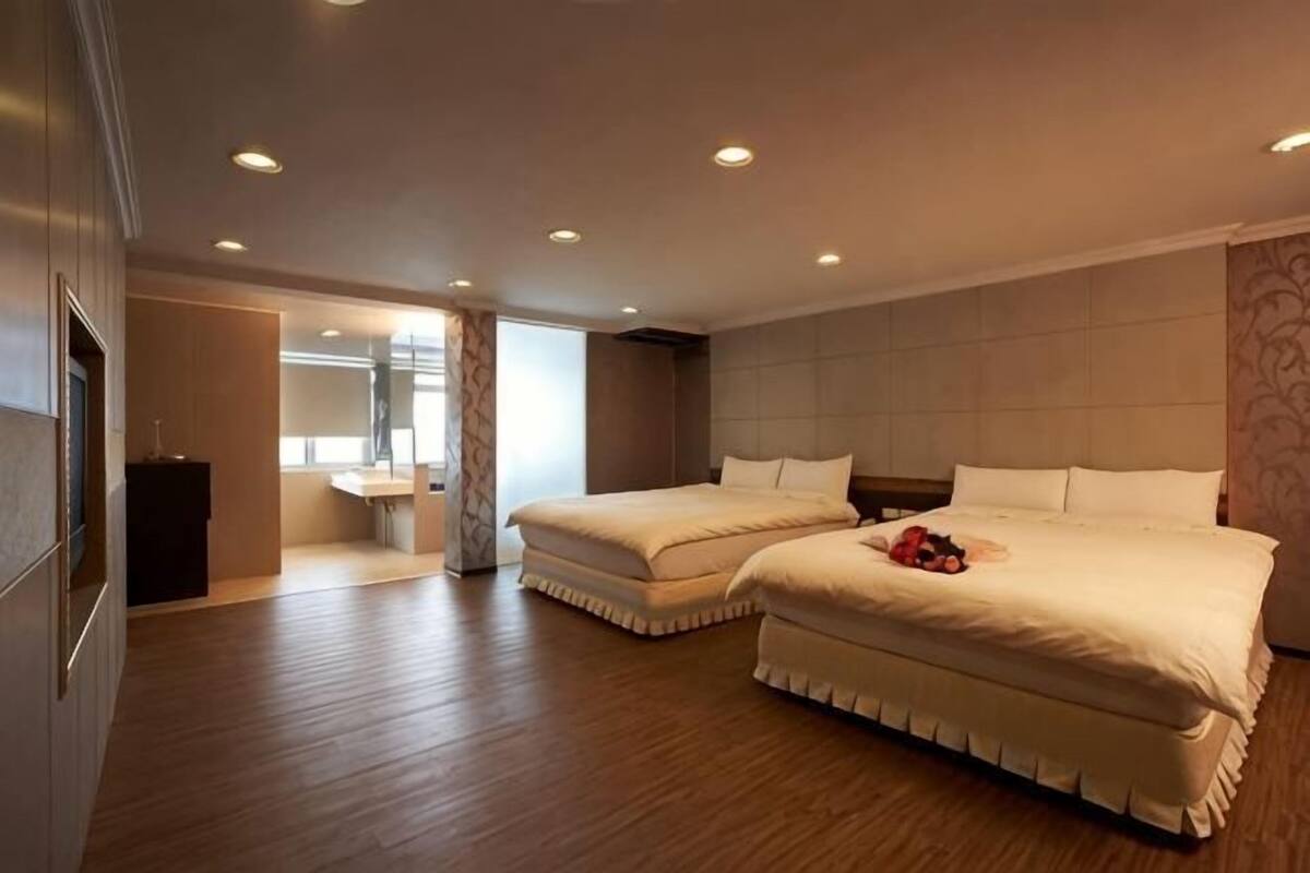 A hotel room with two beds, a wooden floor, a bathroom with a mirror, and a small desk.