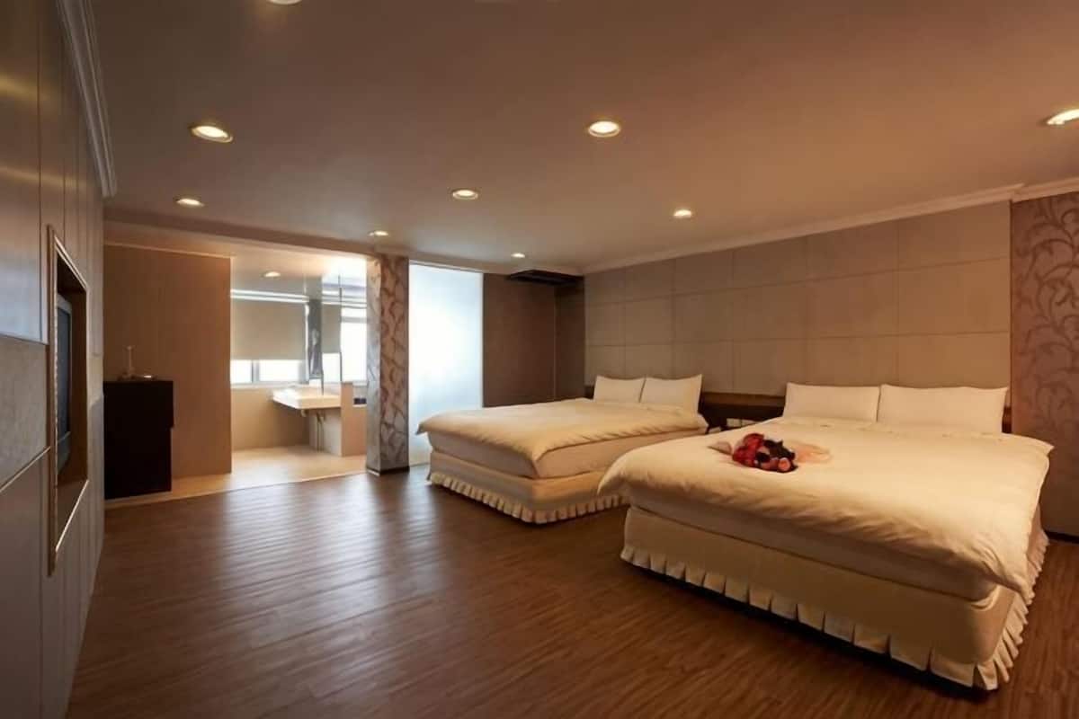 A hotel room with two beds, a wooden floor, a bathroom with a mirror, and a small desk.