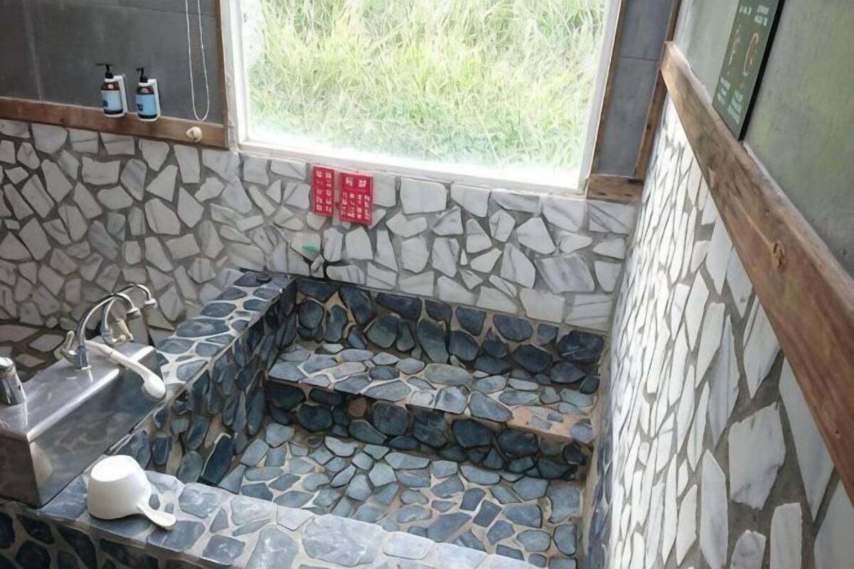 A stone-floored bathroom with a sink, a window, and a red emergency exit sign.