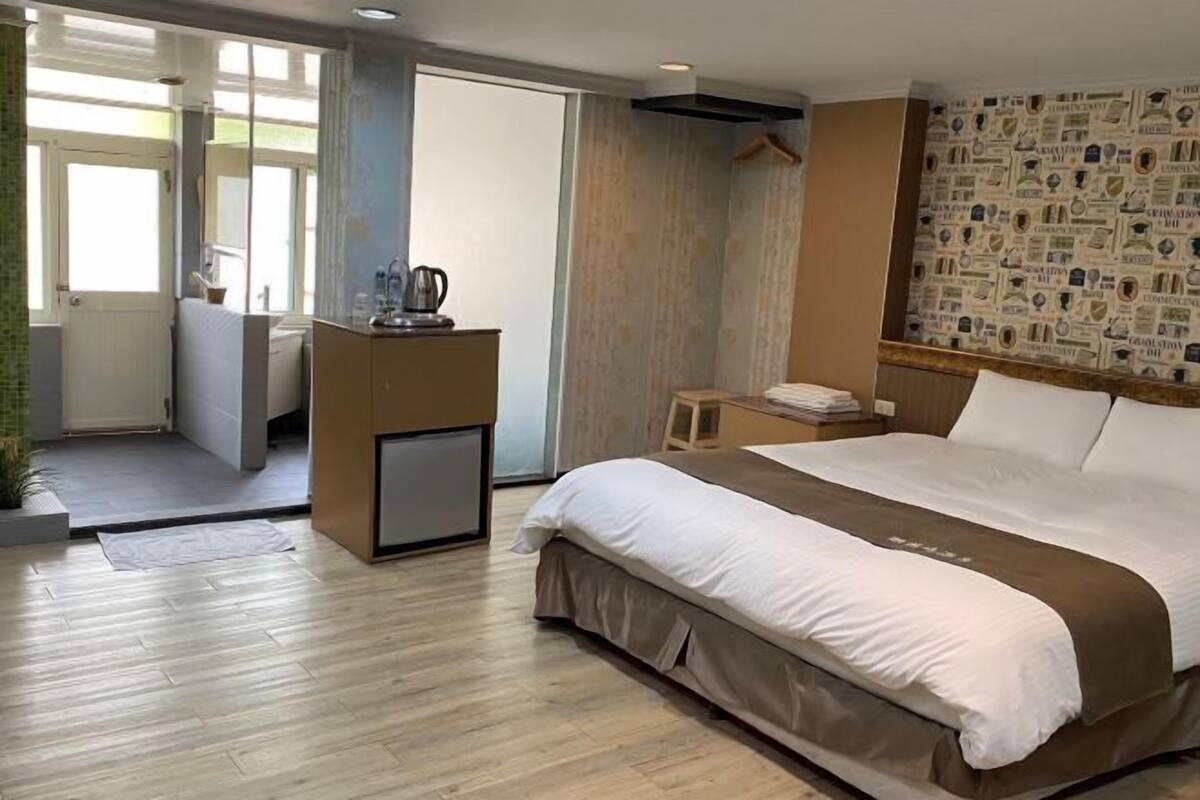 A hotel room with a bed, a desk with a kettle, a small table, and a patterned wall.