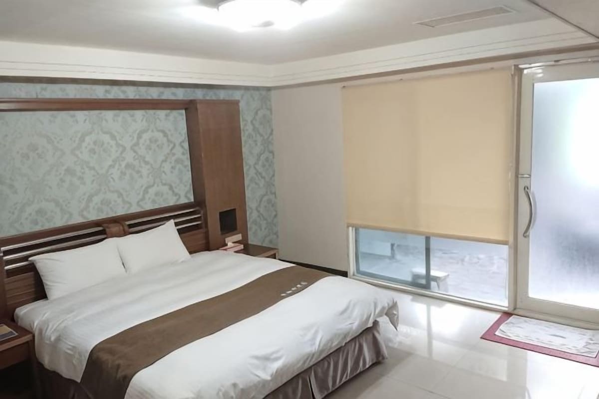Une chambre d’hôtel avec un lit, une table de chevet, une fenêtre donnant sur un paysage agréable et une porte.