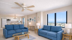 A spacious living room with two blue sofas, a glass coffee table, and a ceiling fan. A kitchen with white cabinets and a dining area with wicker chairs are visible in the background.