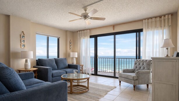 A spacious living room with a large window offering a view of the ocean, a ceiling fan, and a view of the beach through the sliding glass doors.
