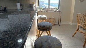 A kitchen with a granite countertop, white cabinets, and a view of the ocean through a large window. There are two stools and a wicker chair in the room.