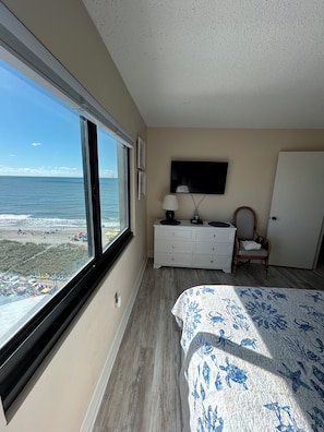 Un dormitorio con un ventanal grande con vistas a la playa, una cama con colcha estampada, un aparador, una silla y un televisor de pantalla plana montado en la pared.