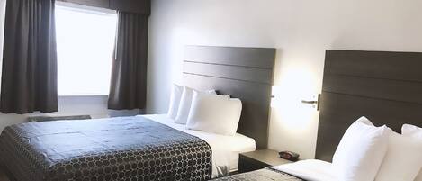 A hotel room with two beds, a large window with curtains, a bedside table with a phone, and a wall-mounted light.