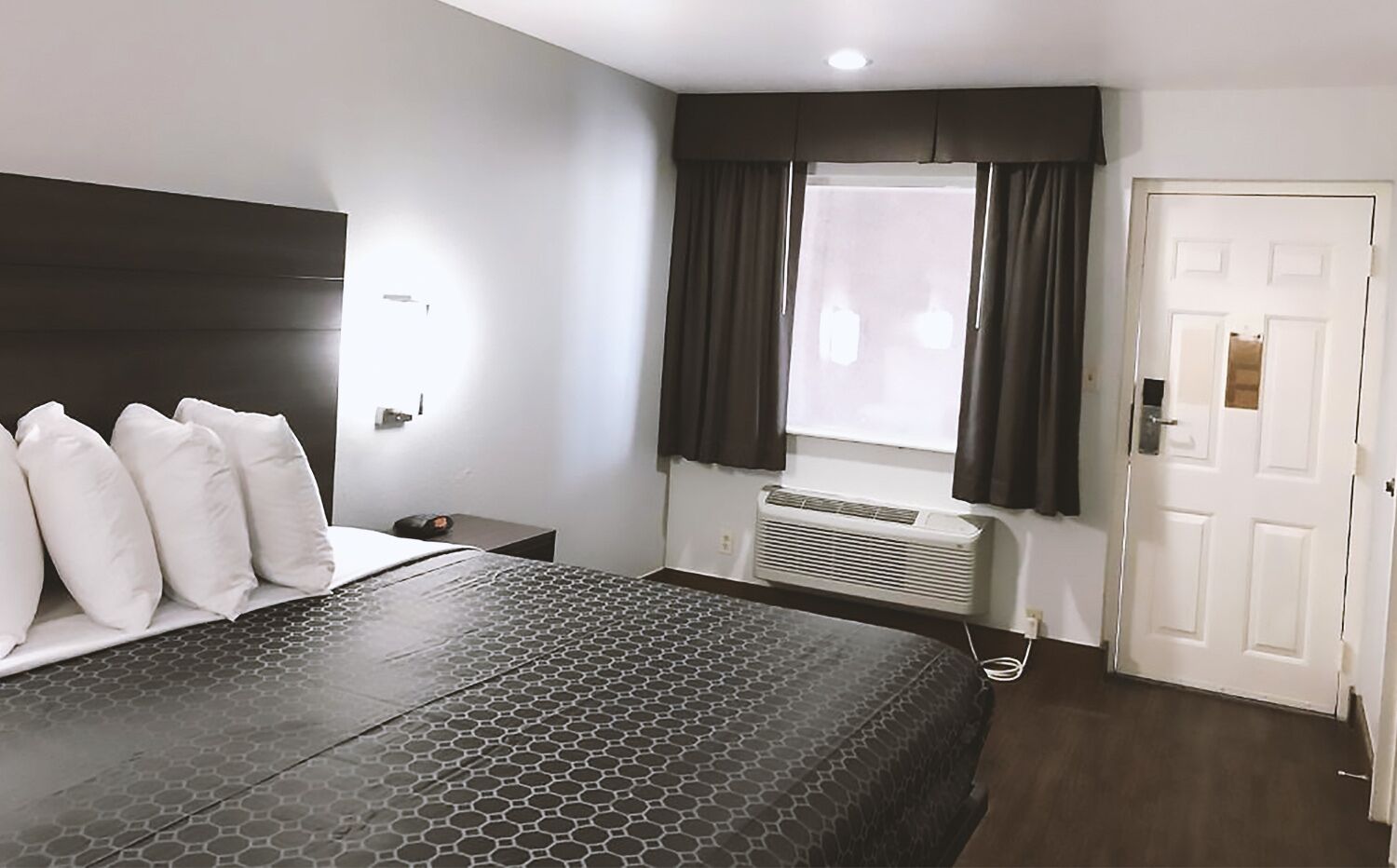 Una camera d'albergo con un letto, un comodino, una finestra con tende, un condizionatore d'aria e una porta.