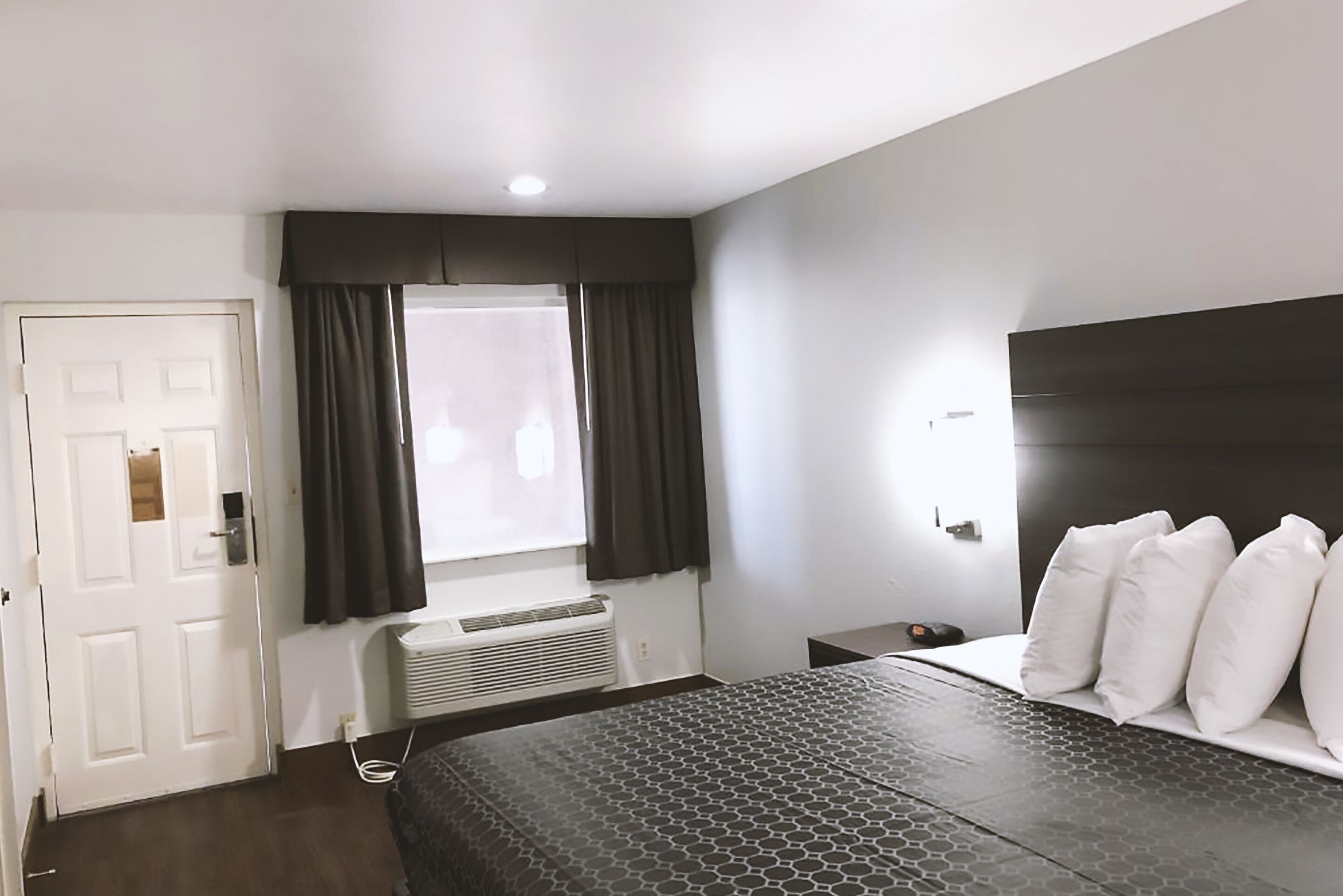 A hotel room with a bed, a bedside table, a window with curtains, a door, and an air conditioning unit.