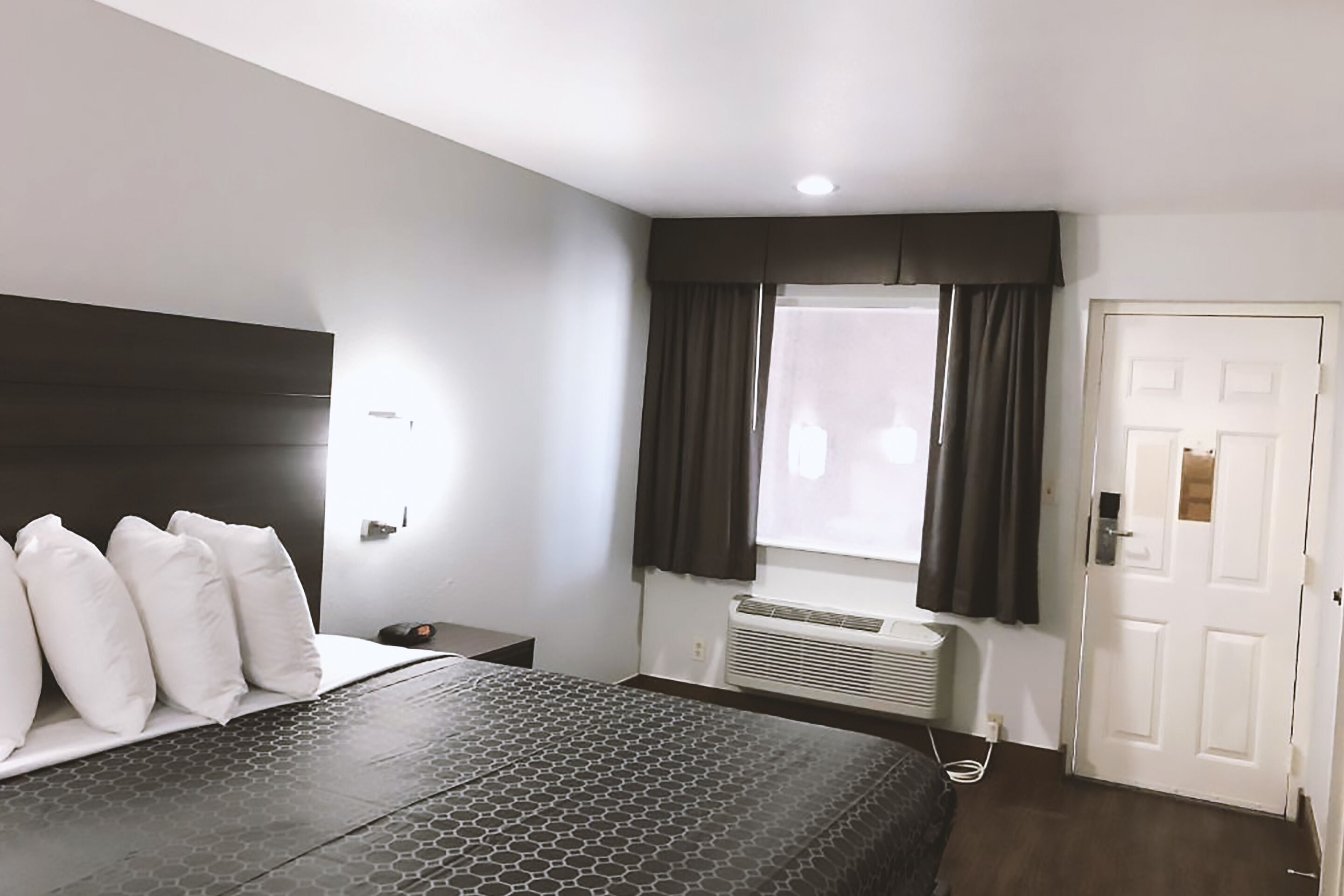 A hotel room with a bed, a nightstand, a window with curtains, a door, and a wall-mounted air conditioning unit.