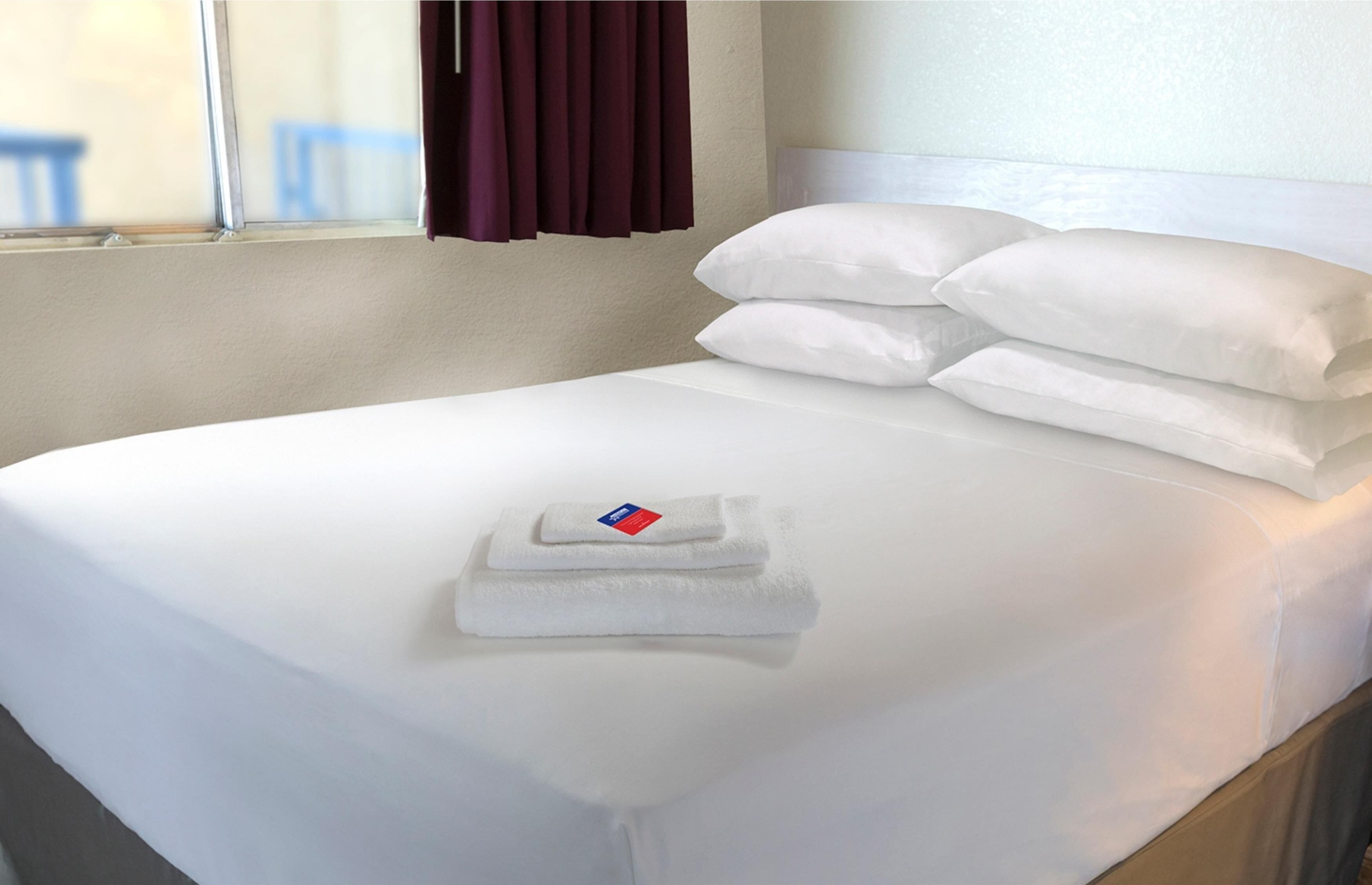 A neatly made bed with white linens and pillows, a folded towel, and a window with curtains.