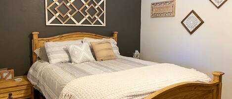 A wooden bed with a white comforter and pillows, a wooden nightstand with a lamp, and decorative wall art.