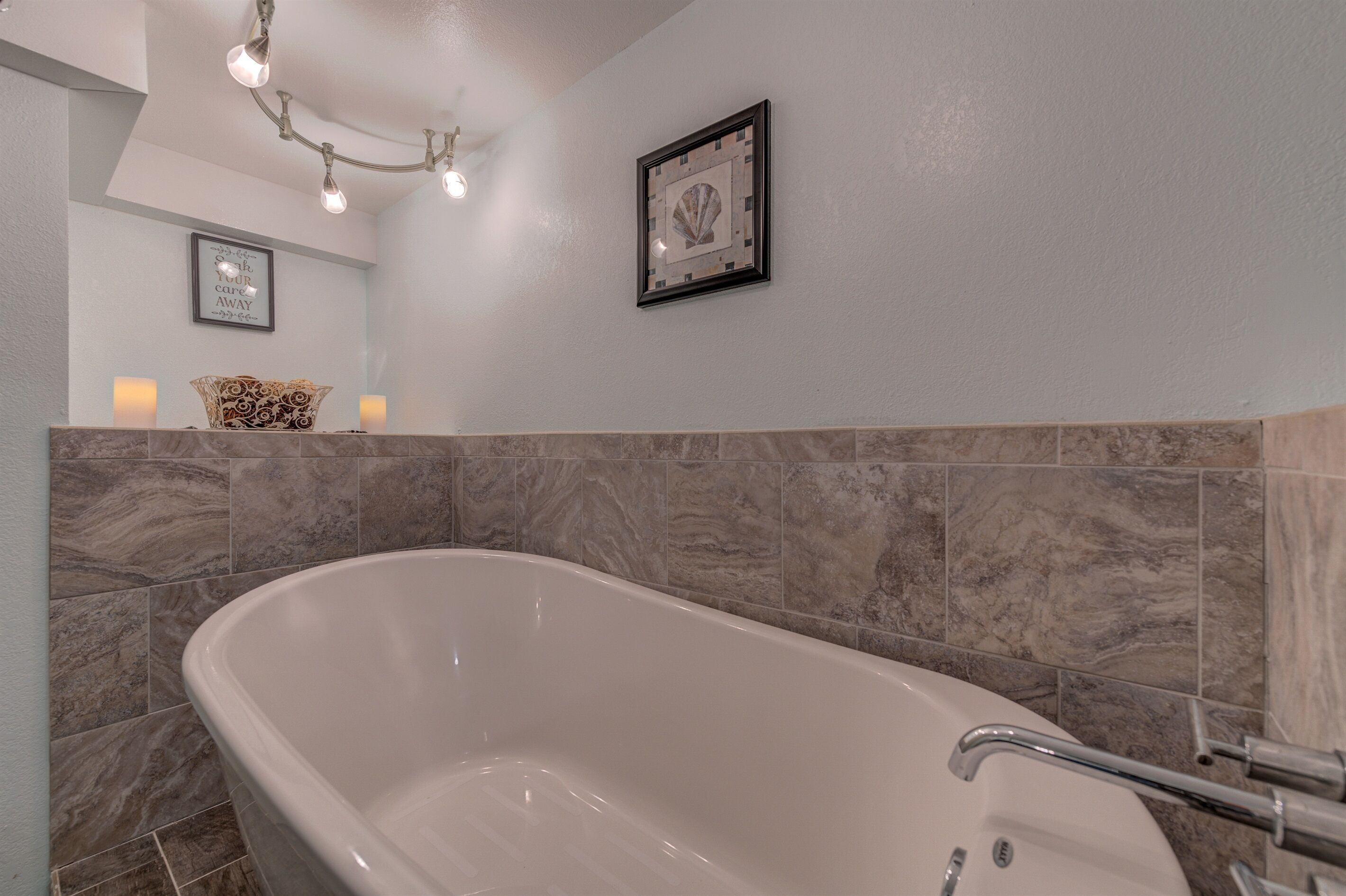A modern bathroom with a freestanding bathtub, marble tiles, a chandelier, and framed artwork on the wall.