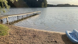 En sandstrand med en træbro, der strækker sig ud i roligt vand, omgivet af træer.