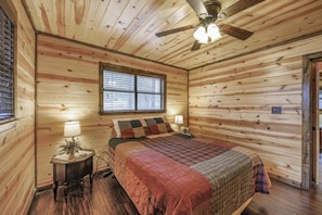 Dormitorio de cabaña de madera con cama, mesita de noche, ventana y ventilador de techo.