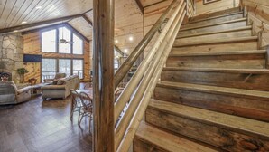 A wooden staircase leading to a lofted area with a stone fireplace and large windows.