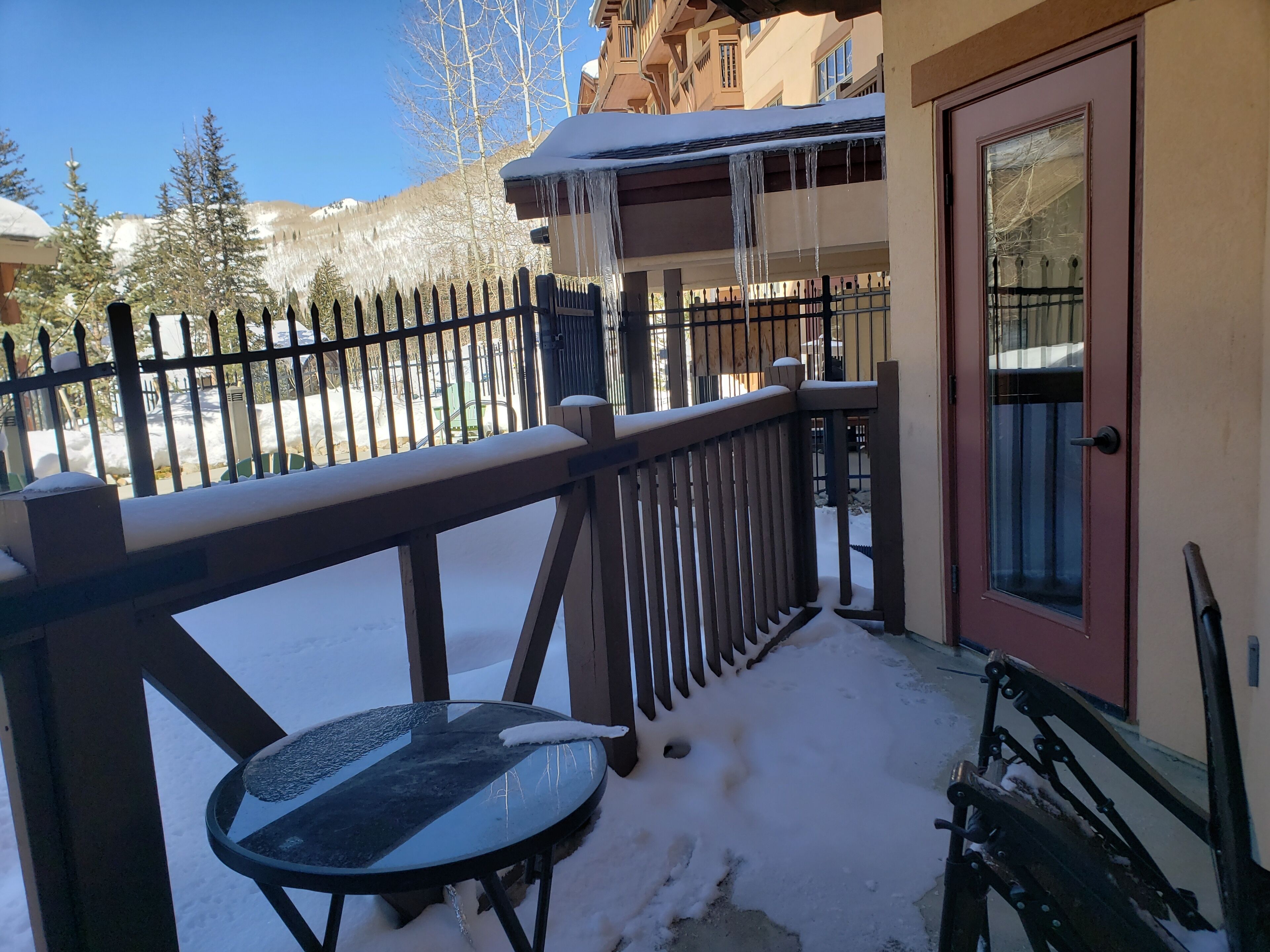 Un balcon avec une table en verre et un banc, entouré d’une clôture en bois et d’un sol recouvert de neige.