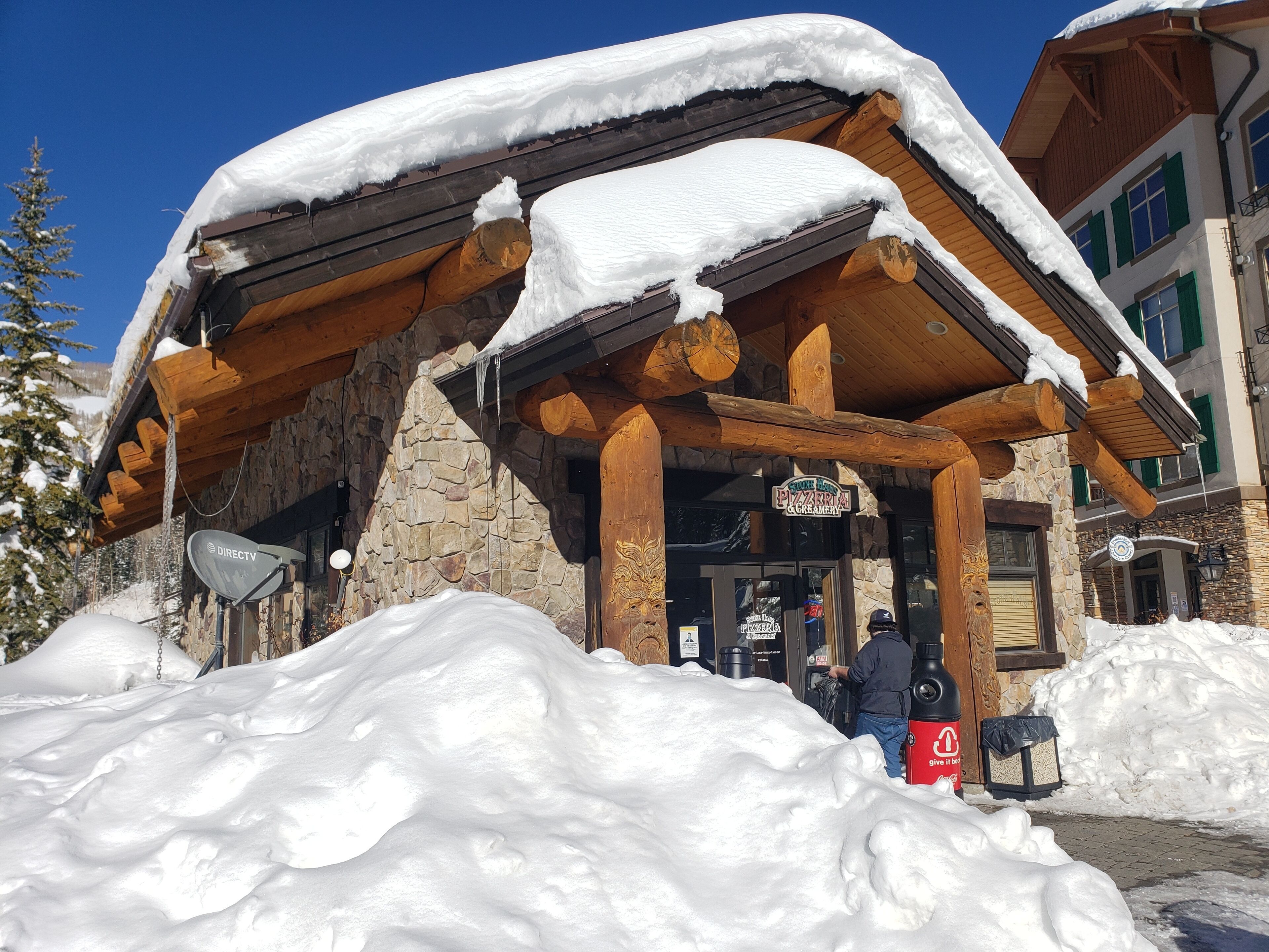 Edificio cubierto de nieve con fachada de piedra y vigas de madera, con un letrero de una pizzería y heladería.