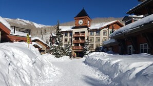 A snow-covered pathway leading to a building with a clock tower, surrounded by snow-laden trees and mountains in the background. - Ski-in / Ski-out best Solitude Resort Village Location (Cottonwood Heights)