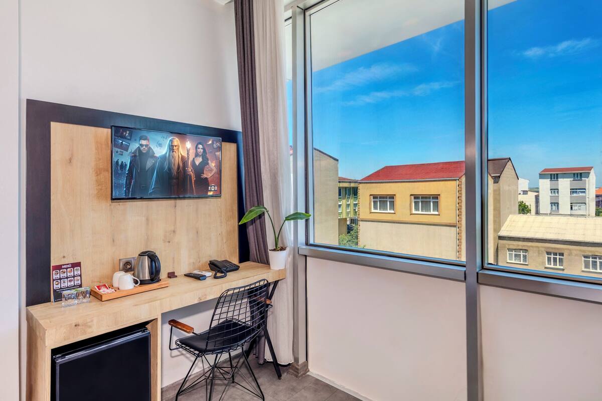 A hotel room with a desk, a TV mounted on the wall, a window with a view of buildings, a plant, and a small refrigerator.