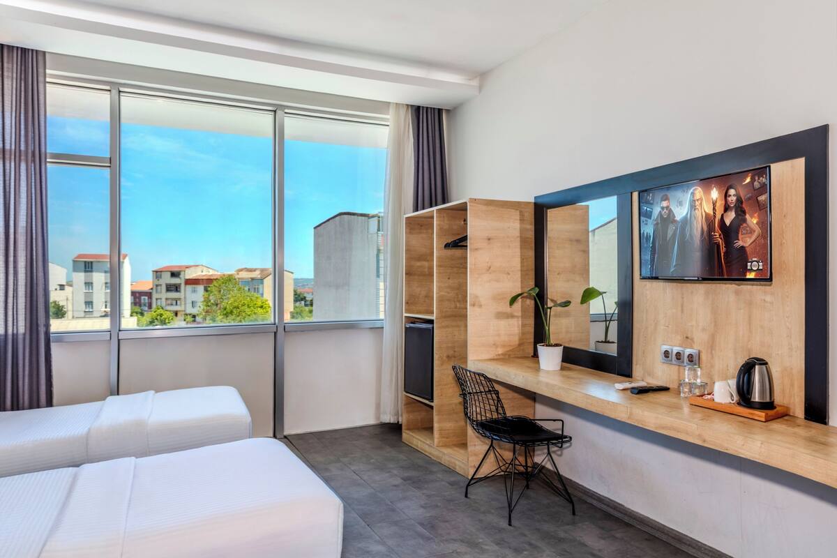 A modern hotel room with a large window, a wooden desk, a chair, a bed, and a TV mounted on the wall.
