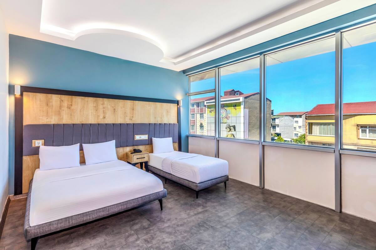 A hotel room with two beds, a wooden headboard, a nightstand, and large windows offering a city view.