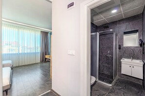 Ein Hotelzimmer mit Dusche, WC und einem kleinen Waschbecken. Ein Bett und ein Fenster mit Vorhängen sind ebenfalls vorhanden.