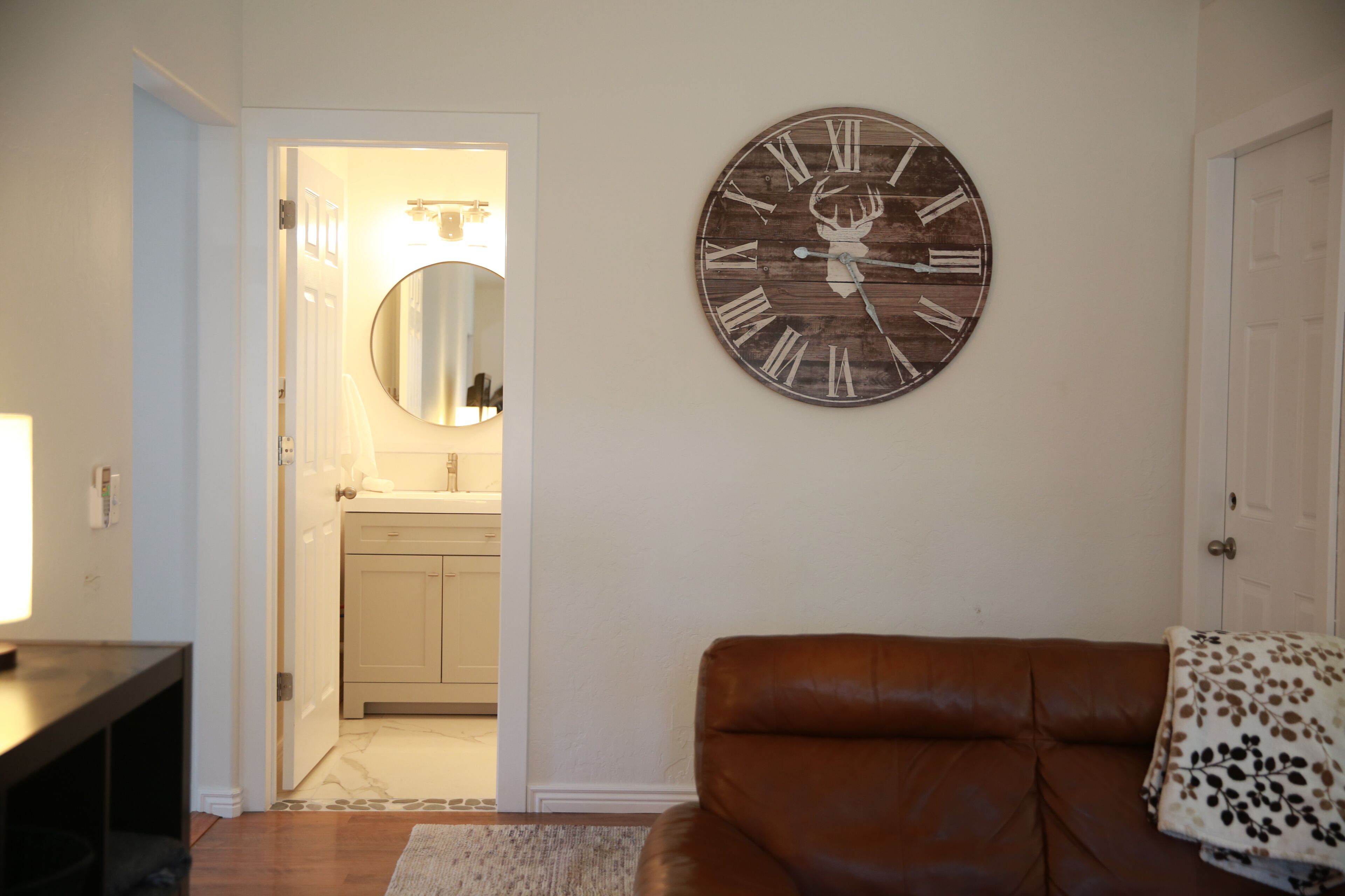 A living room with a brown leather sofa, a wooden clock with a deer head, and a kitchen visible through an open door.