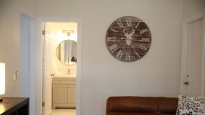 A living room with a brown leather sofa, a wooden clock with a deer head, and a kitchen visible through an open door. - Everything Evergreen (Prescott)
