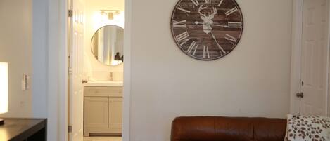 A living room with a brown leather sofa, a wooden clock with a deer head, and a kitchen visible through an open door.