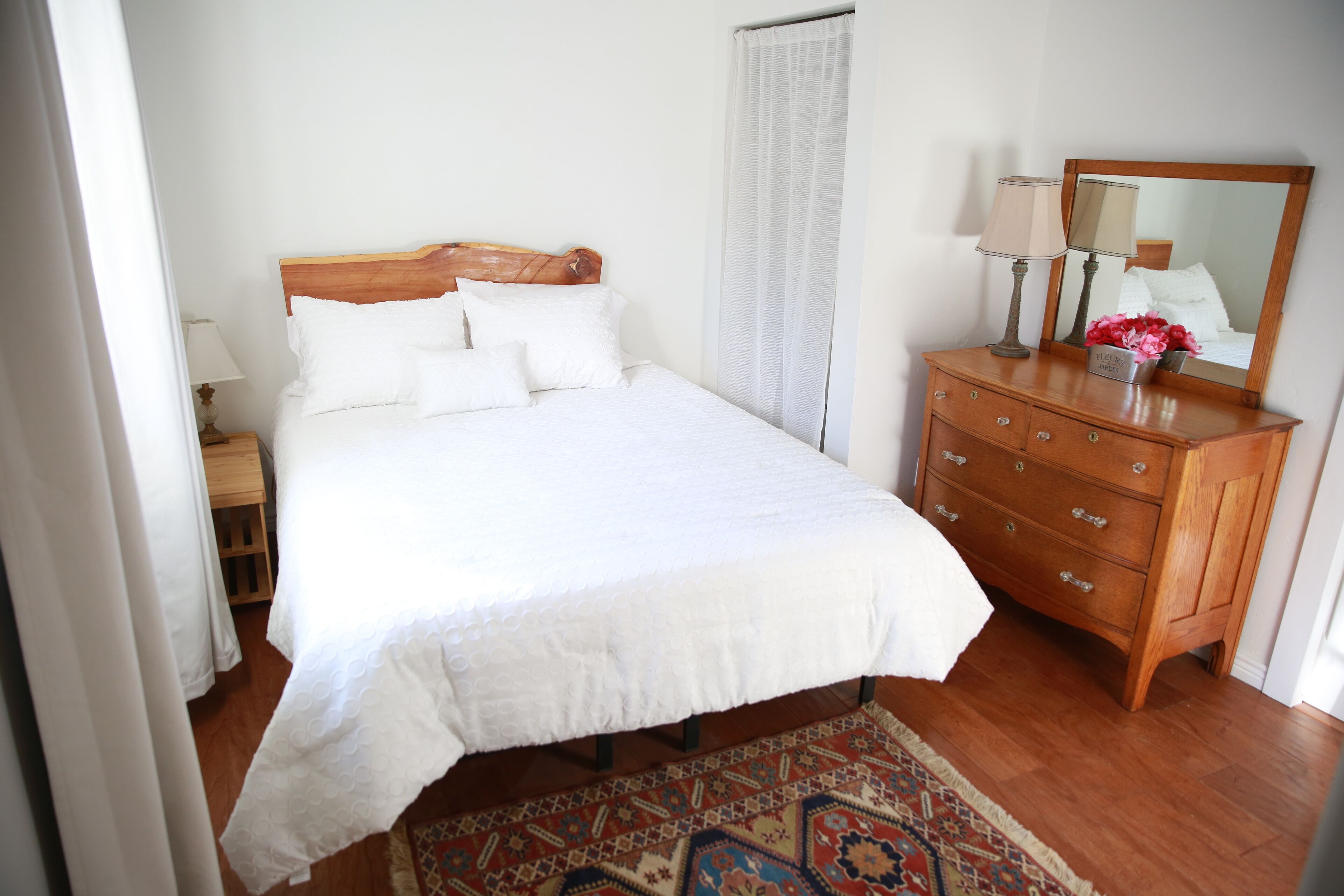 Un lit bien fait, avec une literie blanche, une commode en bois dotée de tiroirs, un miroir et un tapis à motifs posé sur un plancher en bois.