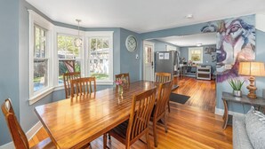 A dining area with a wooden table and chairs, a kitchen in the background, and a large painting on the wall.