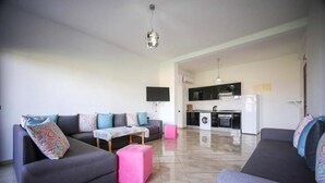 Een moderne woonkamer met twee banken, een salontafel en een roze poef. Achtergrond toont een keuken met zwarte kastjes en witte apparaten.