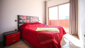 Una camera da letto con una coperta rossa, un comodino rosso e un'ampia finestra con tende.