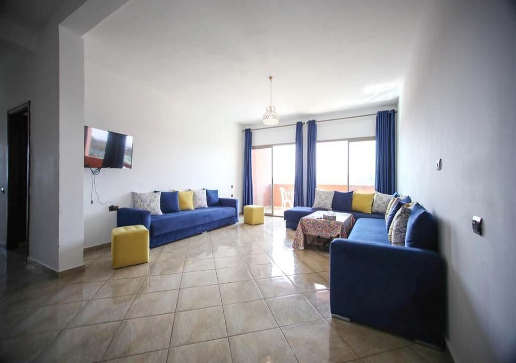 A spacious living room with a blue sofa set, a yellow ottoman, and a table with a floral tablecloth.