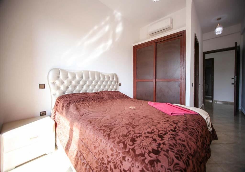 A bedroom with a tufted headboard, a patterned bedspread, a wooden wardrobe, and an air conditioning unit on the wall.