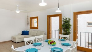 Un coin repas avec une table blanche dressée pour quatre personnes, des assiettes bleues et un saladier de fruits. À l’arrière-plan, on aperçoit un canapé blanc avec des coussins bleus, et à droite, une plante en pot.