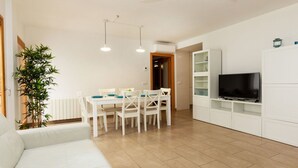 Un salon moderne avec un canapé blanc, une table à manger avec des chaises, un téléviseur posé sur un meuble blanc et une grande plante.