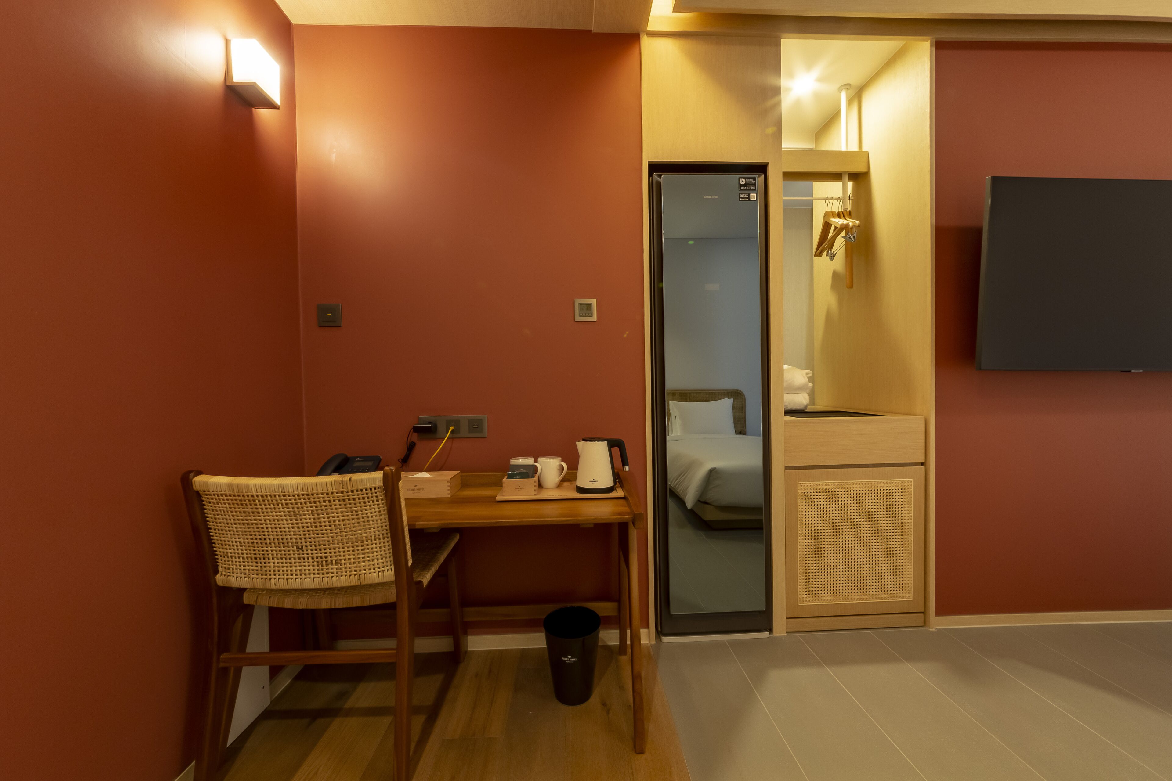 A hotel room with a wooden desk, chair, and a small trash bin. There is a bed with a white mattress and a wooden headboard. A flat-screen TV is mounted on the wall.