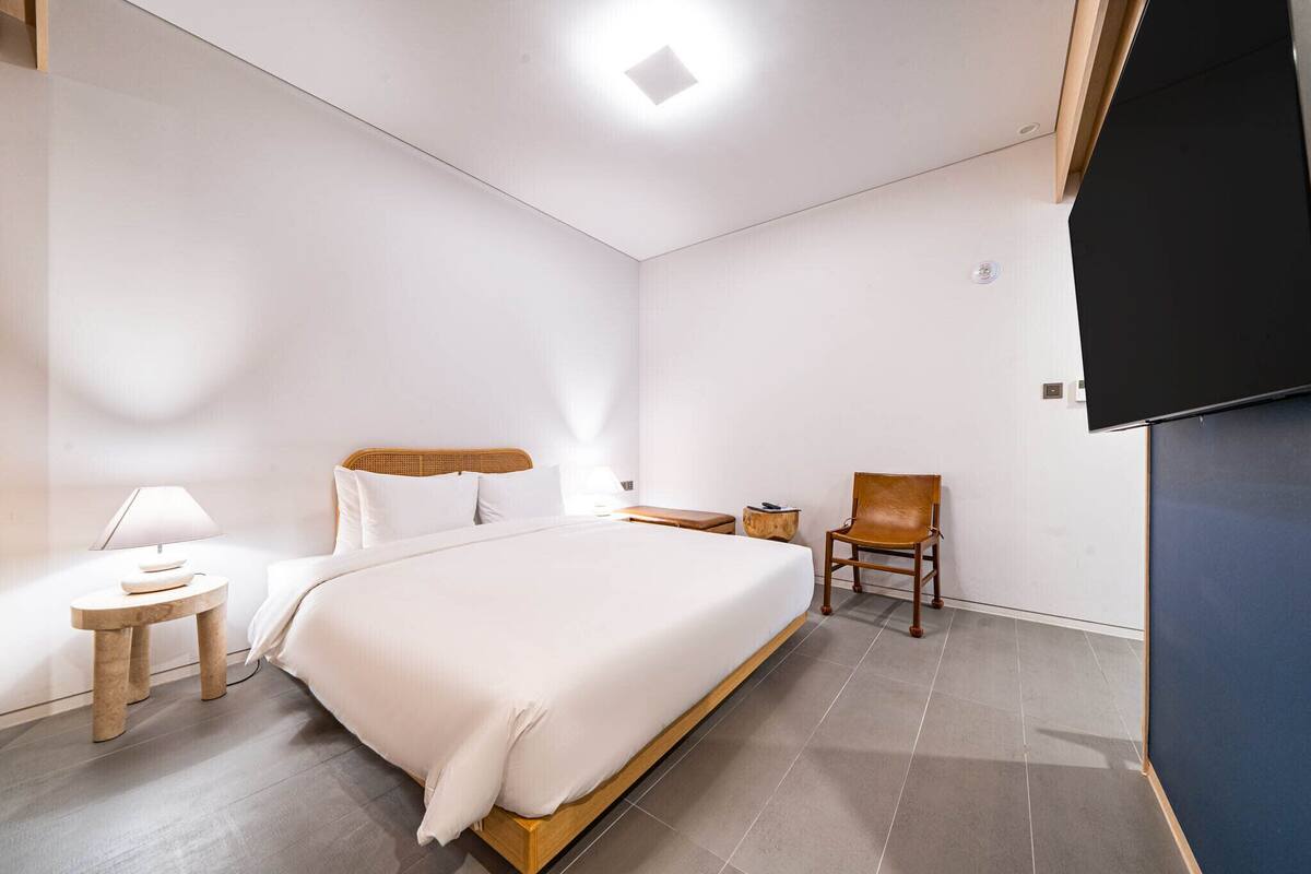 A modern hotel room with a large bed, a wooden headboard, a small bedside table with a lamp, a chair, and a flat-screen TV mounted on the wall.