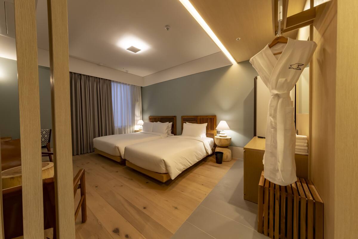 A hotel room with two beds, a wooden floor, a desk with chairs, a bedside table with a lamp, and a robe hanging on a wooden stand.