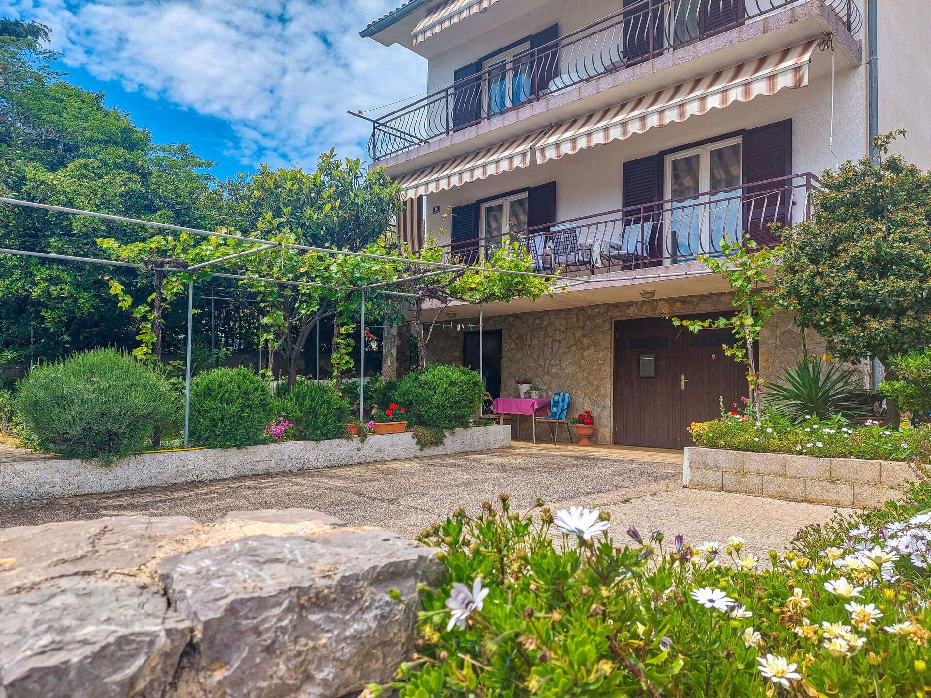 A two-story building with a balcony, surrounded by greenery and flowers.