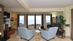 A spacious living room with a large window offering a view of the sea, two grey armchairs, a small round table, and a television mounted on a brick wall.