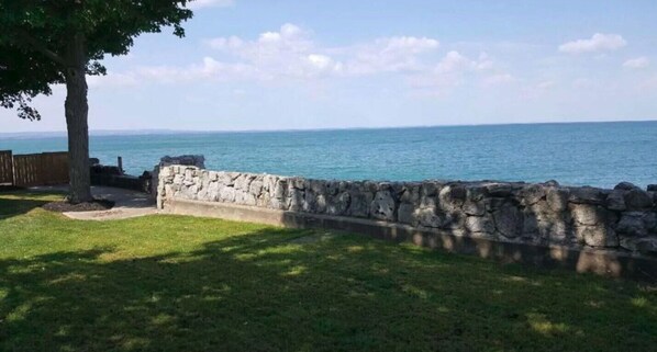 A stone wall with a grassy area and a tree in the foreground, leading to a view of the ocean.