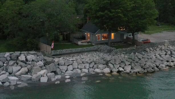 A lakeside cabin with a stone wall, surrounded by trees and a dock.