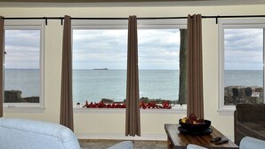 A room with a view of the sea, featuring a large window with curtains, a seating area with a table, and a small plant on the windowsill.