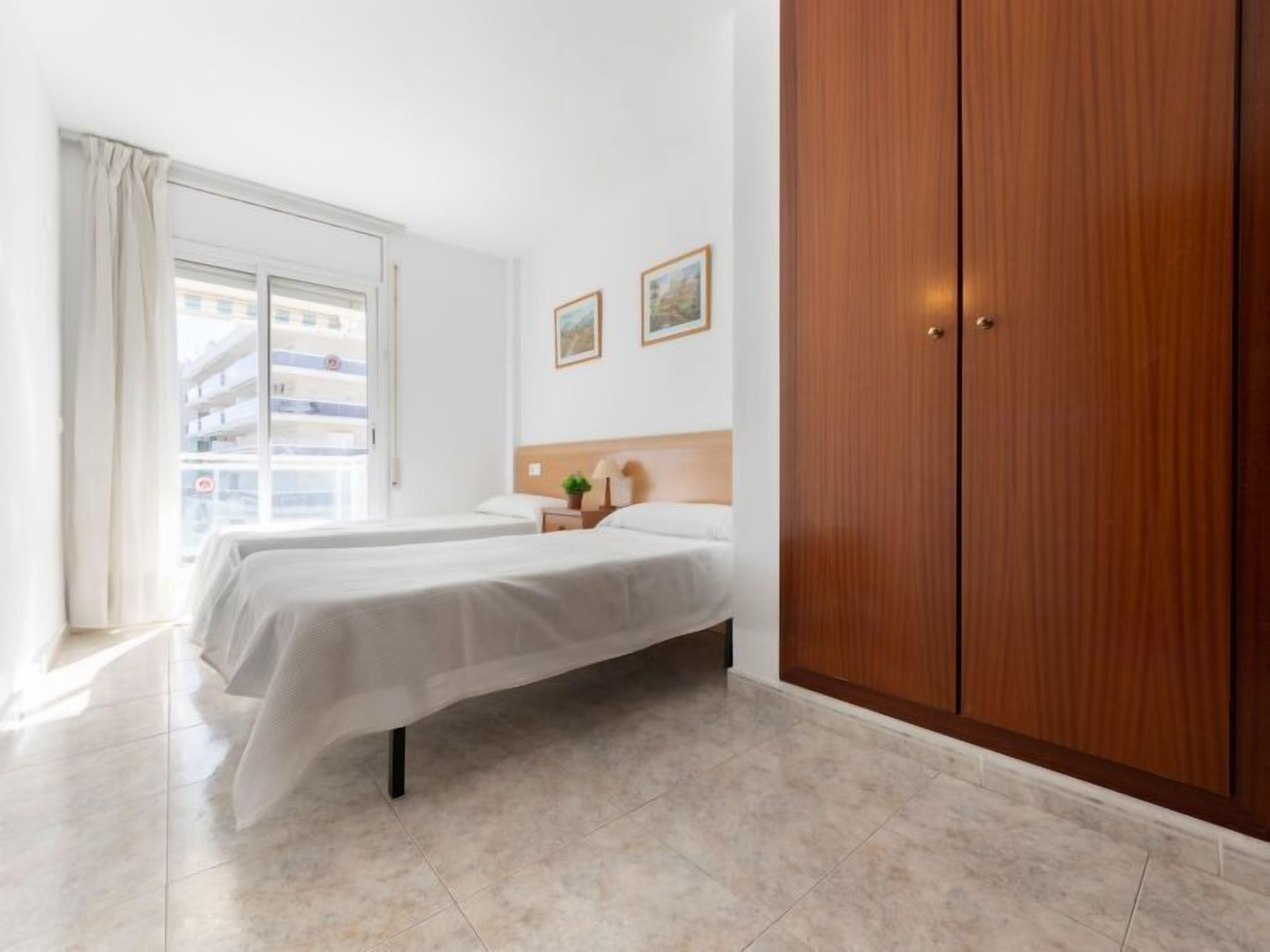 A hotel room with two beds, a wooden wardrobe, a window with curtains, and a view of a building outside.