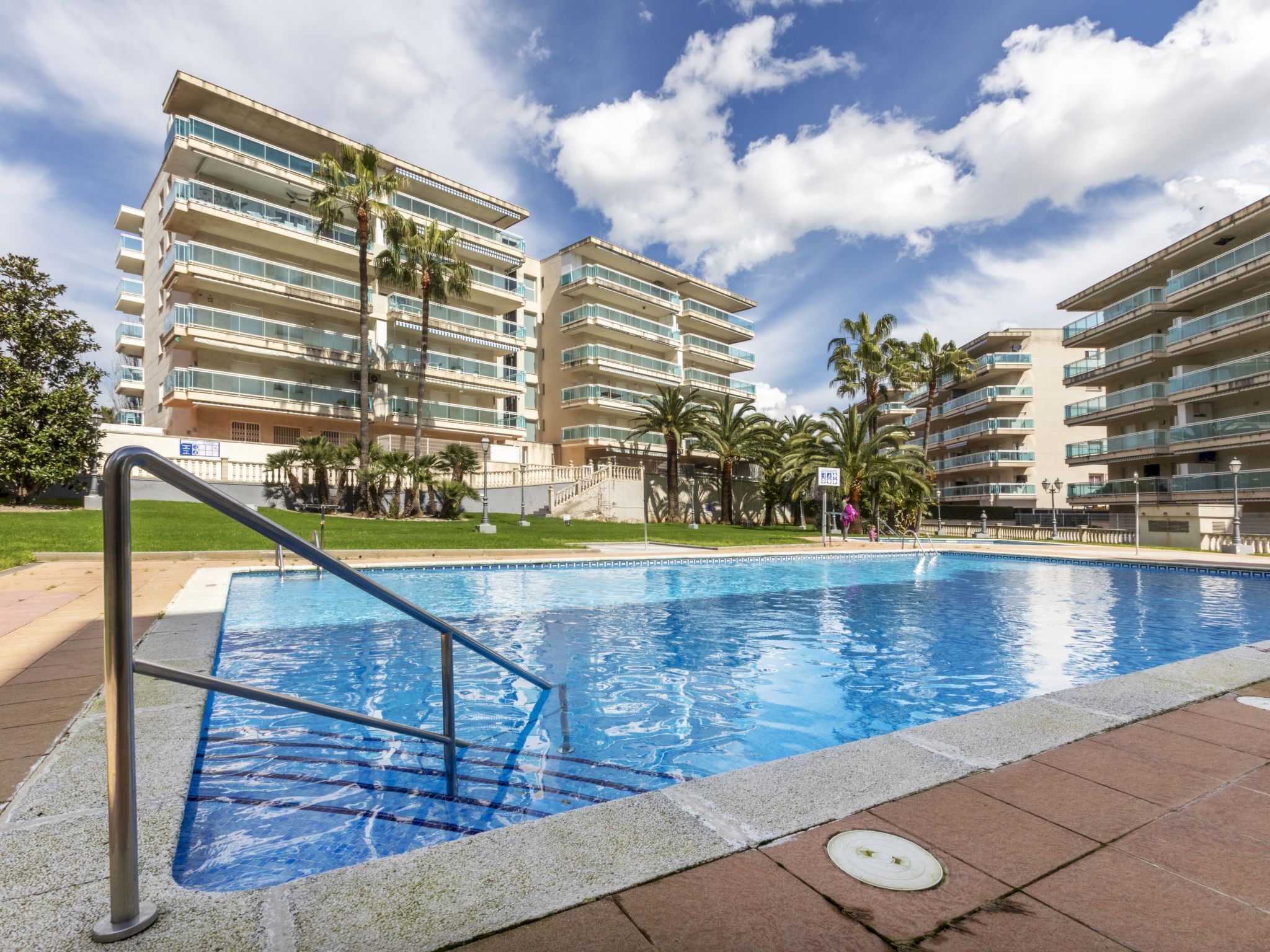 A swimming pool with a metal ladder, surrounded by palm trees and a multi-story building with balconies.