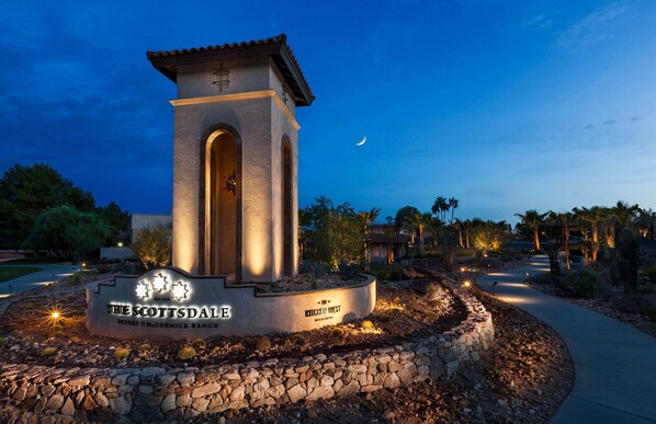 A stone monument with a cross, surrounded by a landscaped garden with cacti and palm trees, and a crescent moon in the sky.